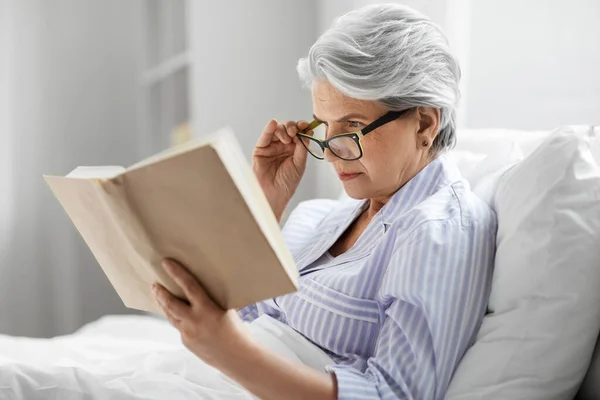 Anciana en gafas leyendo libro en la cama en casa — Foto de Stock