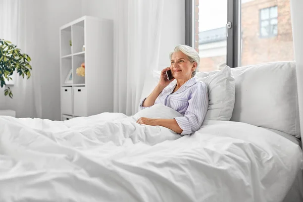 Mulher sênior chamando no smartphone na cama em casa — Fotografia de Stock