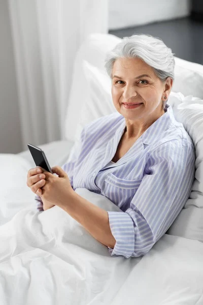 Mulher sênior feliz usando smartphone na cama em casa — Fotografia de Stock