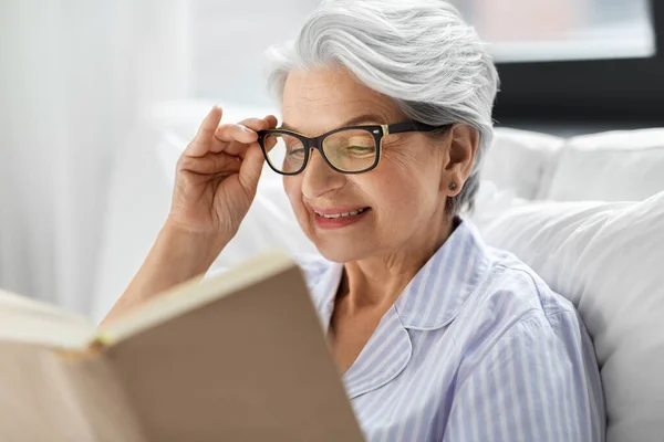 Gözlüklü yaşlı kadın evde yatakta kitap okuyor. — Stok fotoğraf