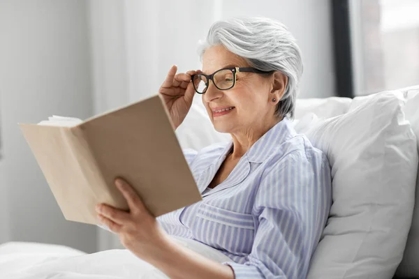 Старуха в очках читает книгу в постели дома — стоковое фото