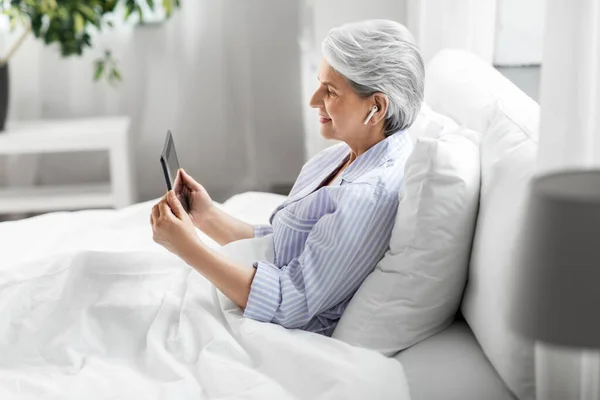 Donna anziana con tablet pc e auricolari a letto — Foto Stock