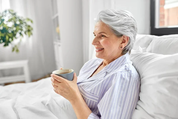 Ηλικιωμένη γυναίκα με φλιτζάνι καφέ στο κρεβάτι στο σπίτι — Φωτογραφία Αρχείου