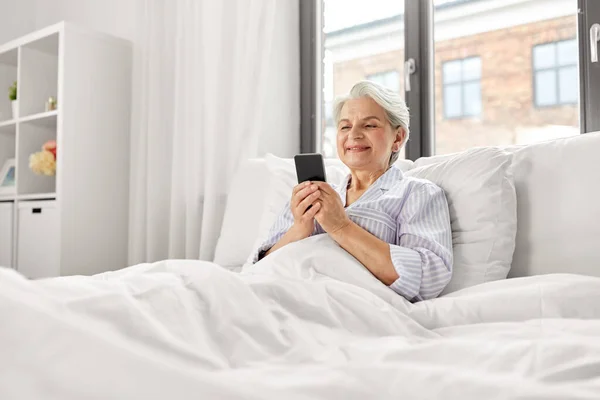 Mujer mayor feliz usando el teléfono inteligente en la cama en casa —  Fotos de Stock