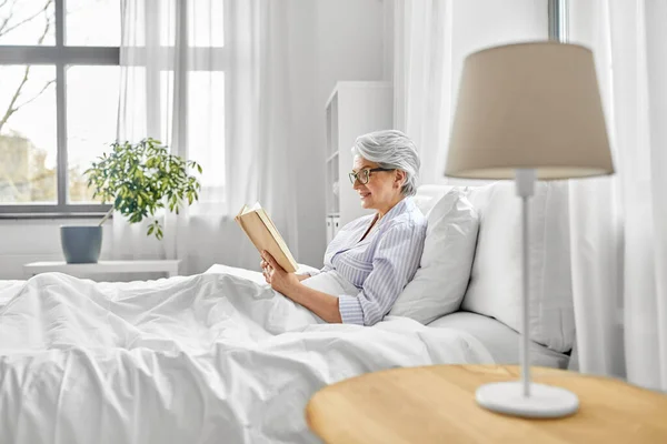 Mulher velha em óculos livro de leitura na cama em casa — Fotografia de Stock