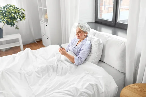 Mulher idosa com smartphone e fones de ouvido na cama — Fotografia de Stock