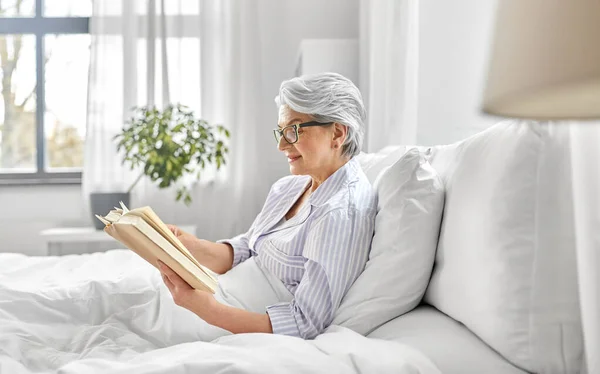 Old woman in glasses reading book in bed at home — Stock Photo, Image