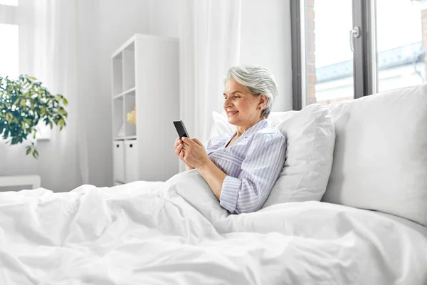 Mujer mayor feliz usando el teléfono inteligente en la cama en casa —  Fotos de Stock