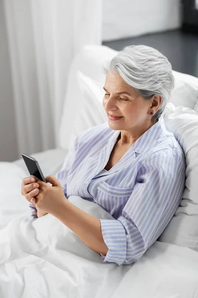 Happy senior woman using smartphone in bed at home — Stock Photo, Image