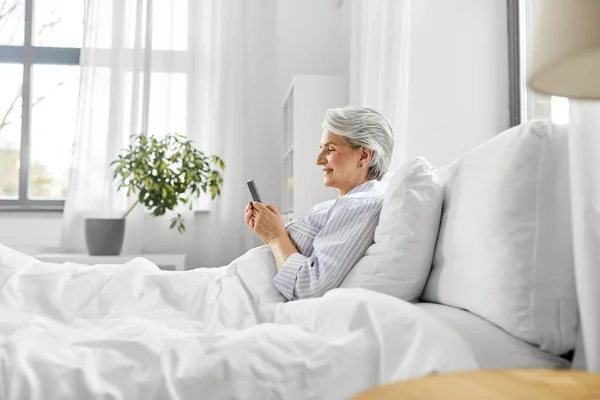 Glückliche Seniorin nutzt Smartphone zu Hause im Bett — Stockfoto