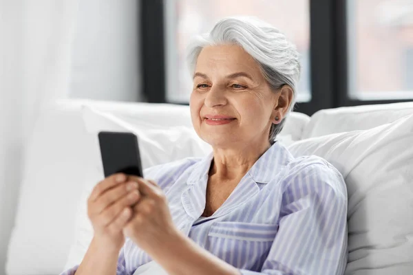 Szczęśliwy senior kobieta za pomocą smartfona w łóżku w domu — Zdjęcie stockowe