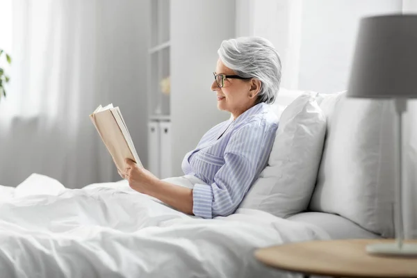 Old woman in glasses reading book in bed at home — Stock Photo, Image