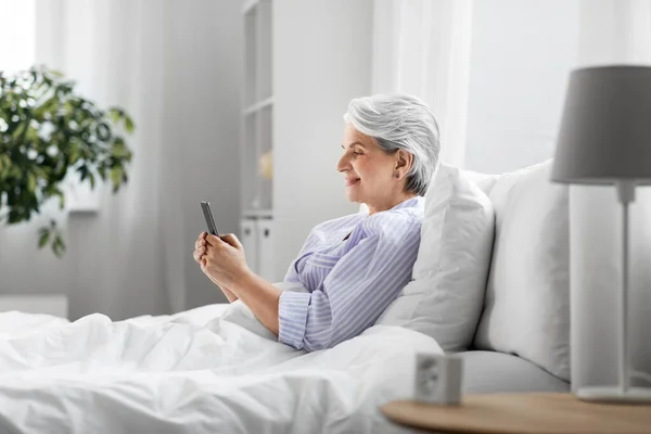 Happy senior woman using smartphone in bed at home — Stock Photo, Image