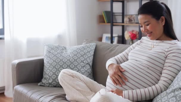 Счастливая беременная азиатка сидит дома на диване — стоковое видео