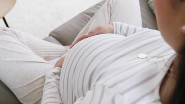 Счастливая беременная азиатка сидит дома на диване — стоковое видео