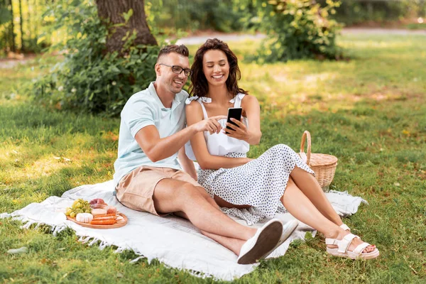 Pareja feliz con teléfono inteligente en el picnic en el parque —  Fotos de Stock