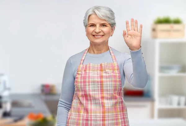 Улыбающаяся пожилая женщина в кухонном фартуке машет рукой — стоковое фото