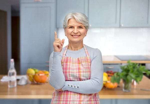 Sonriente mujer mayor en delantal apuntando con el dedo hacia arriba — Foto de Stock