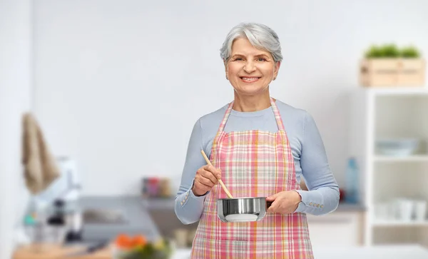 Stará žena v kuchyni zástěra s hrncem vaření potravin — Stock fotografie