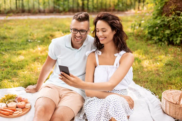 Pareja feliz con teléfono inteligente en el picnic en el parque —  Fotos de Stock