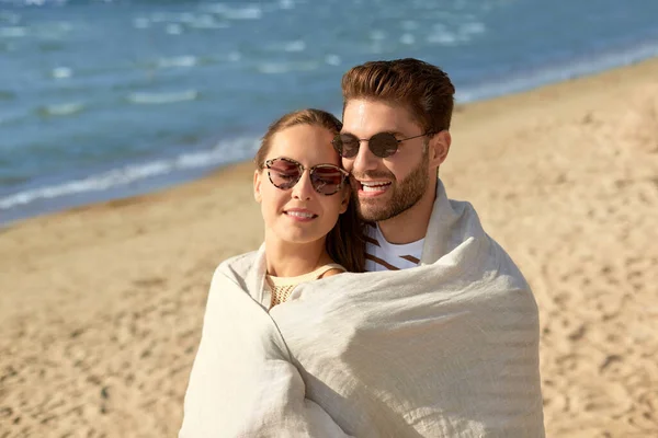 Feliz casal coberto com cobertor abraço na praia — Fotografia de Stock