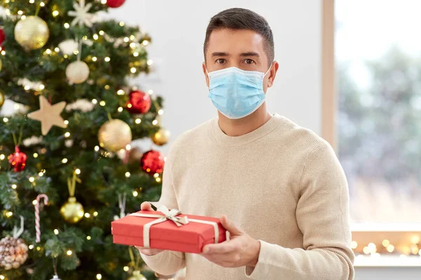 Homem em máscara com presente na árvore de natal em casa — Fotografia de Stock