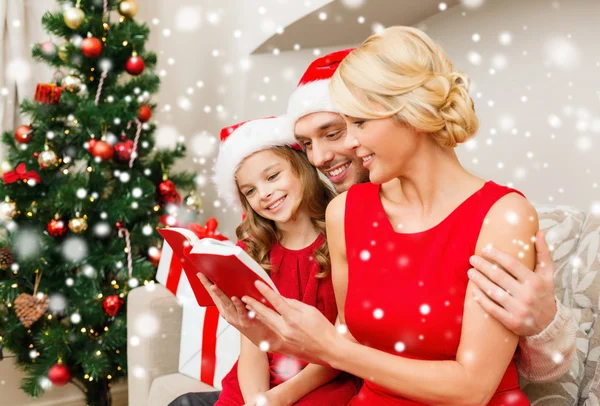 Smiling family in santa hats reading book at home — Stock Photo, Image