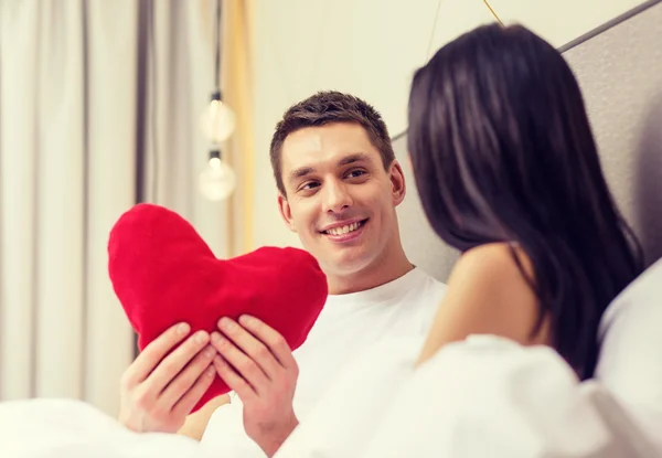 Smiling couple in bed with red heart shape pillow — Stock Photo, Image