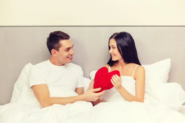 Улыбающаяся пара в постели с красной подушкой в форме сердца — стоковое фото