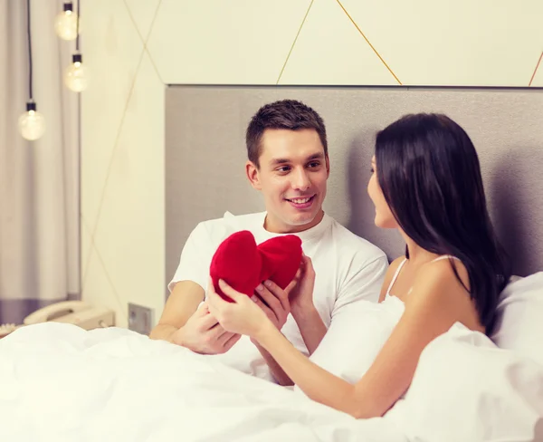 Casal sorridente na cama com travesseiro em forma de coração vermelho — Fotografia de Stock