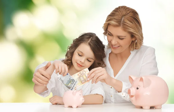 Mother and daughter putting money to piggy banks Stock Photo