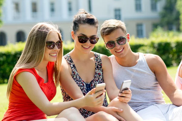 Amigos sonrientes con teléfonos inteligentes sentados en el parque —  Fotos de Stock