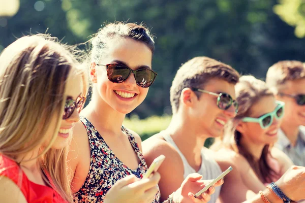 Smiling friends with smartphones sitting in park — Stock Photo, Image