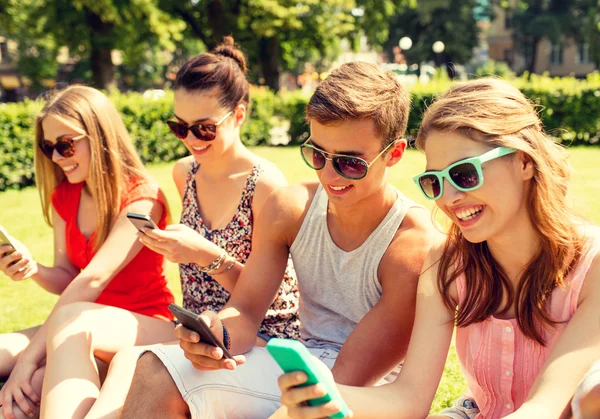 Smiling friends with smartphones sitting on grass — Stock Photo, Image
