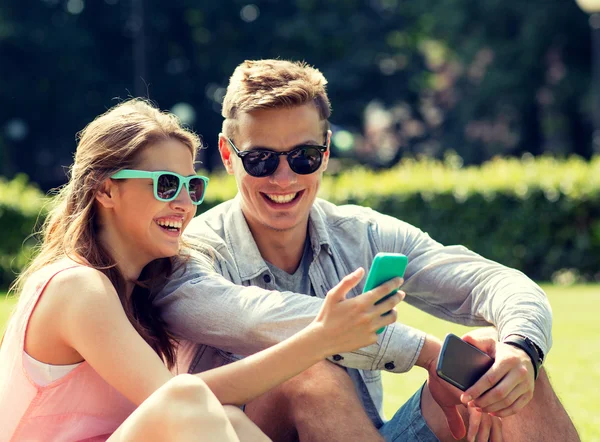 Smiling friends with smartphones sitting in park — Stock Photo, Image