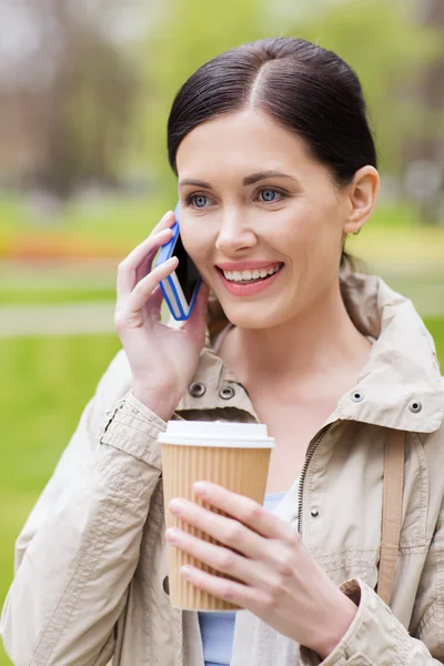 Mujer sonriente con teléfono inteligente y café en el parque —  Fotos de Stock