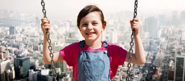 Happy little girl swinging on swing over city — Stok fotoğraf
