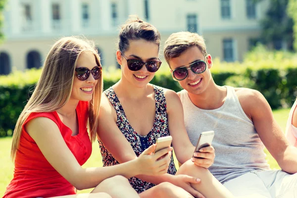 Amigos sonrientes con teléfonos inteligentes sentados en el parque — Foto de Stock