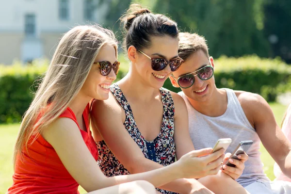 Улыбающиеся друзья со смартфонами, сидящими в парке — стоковое фото