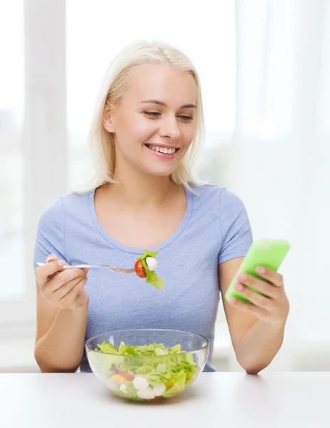 Mulher sorridente com smartphone comendo salada em casa — Fotografia de Stock