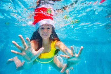 Mutlu küçük kız suya dalıyor ve yüzüyor. Havuzda Noel Baba şapkası takıyor ve elleriyle el sallıyor.