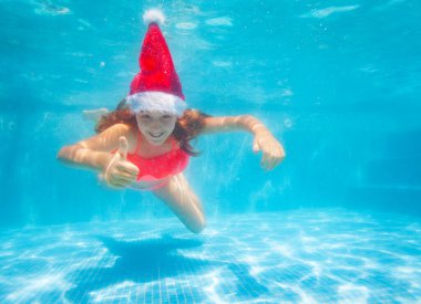 Havuzda Noel Baba şapkası takmış, el hareketleriyle yüzen mutlu bir kız.