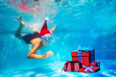 Noel Baba şapkasıyla havuza dalan maskeli mutlu çocuk Noel hediyesi kutularına uzanıyor.