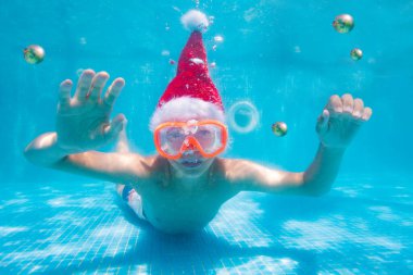 Skuba maskeli yakışıklı çocuk havuzda Noel süsleri takan Noel Baba şapkası takmış ve su altında yüzüyor.