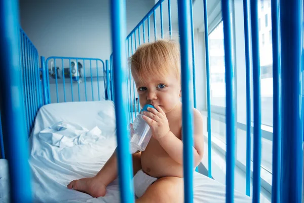 小さな幼児は大きな金属フレームで病院のベッドに座って ボトルからミルクを飲むカメラで見る — ストック写真