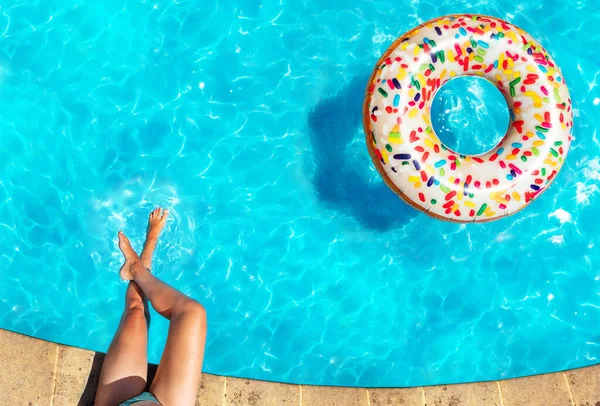 边界上的女人的腿和充气的糖果甜甜圈浮标在游泳池里游来游去 — 图库照片