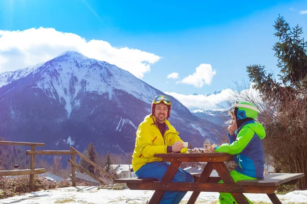 父亲穿着滑雪服和小男孩坐在一起 一边滑雪一边欣赏高山美景 一边吃午饭 — 图库照片