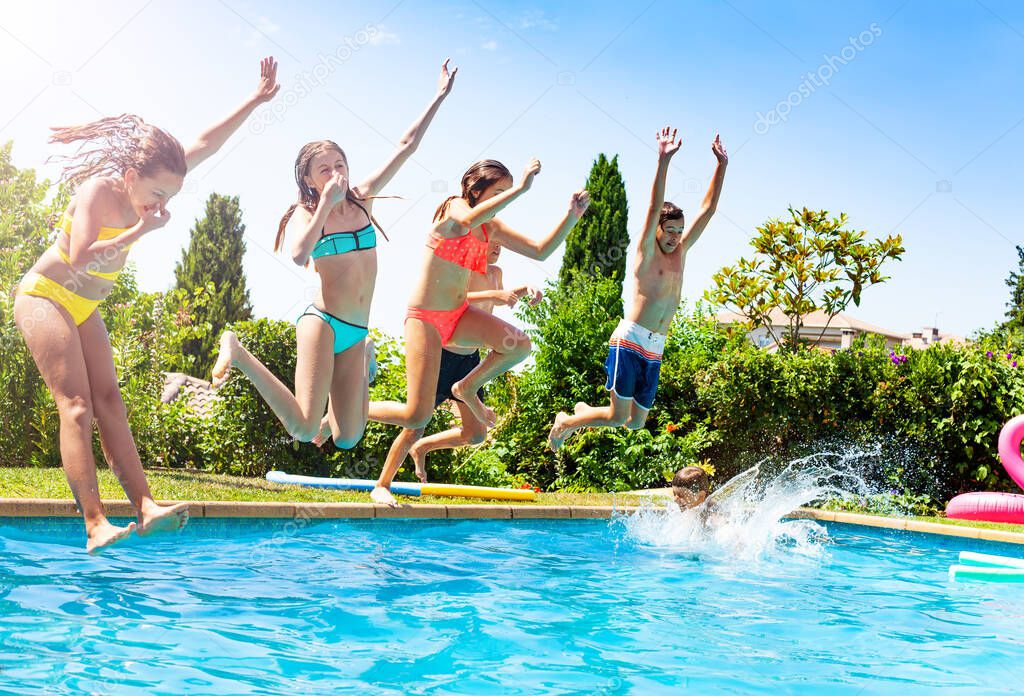 Happy kids in a group of friends jump into water of swimming pool together splashing down and lifting hands