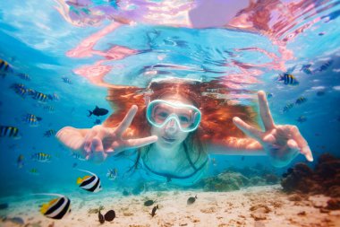 Kızın dalış maskeli portresi balık okulları arasında su altında yüzüyor ve zafer jestini elleriyle gösteriyor.