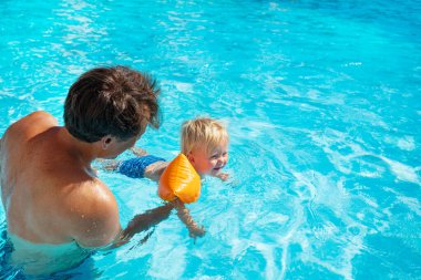 Bir babanın yukarıdan görünüşü oğluna havuzda yüzmeyi öğretiyor şişme omuz kemerleriyle.